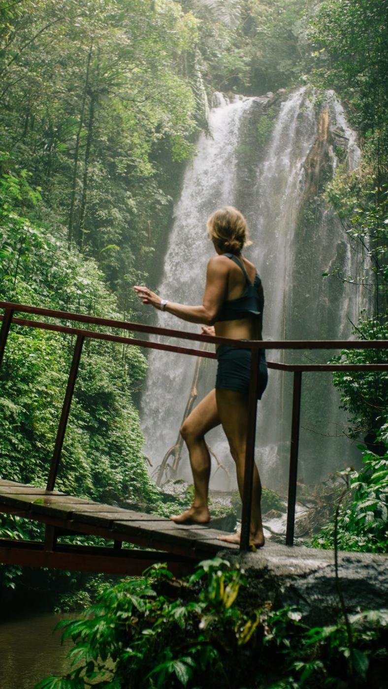 A pilgrim on a wooden bridge facing a jungle waterfall